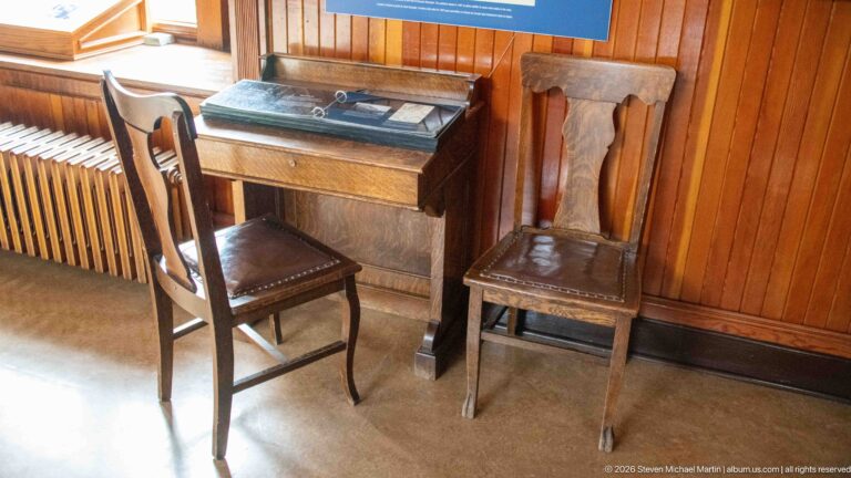 Small wooden desk with a glass-covered top display and two chairs against varnished wall paneling.