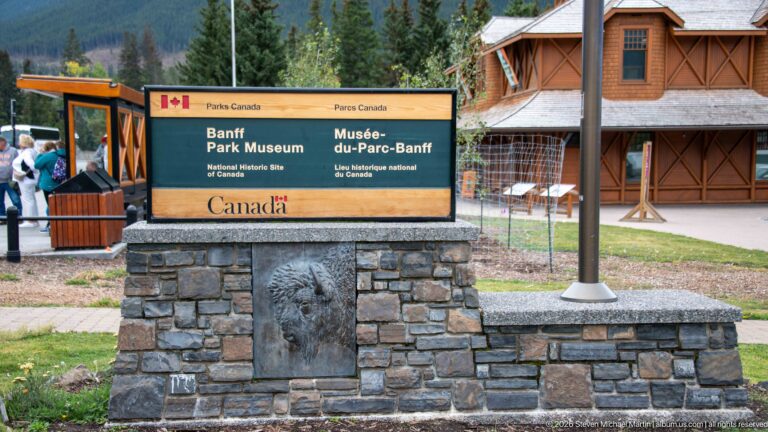 Bilingual Banff Park Museum sign on a stone base, with the brown timber museum building and trees behind.