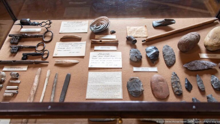 Display case with stone points, blades, polished objects, metal implements, and small descriptive labels.