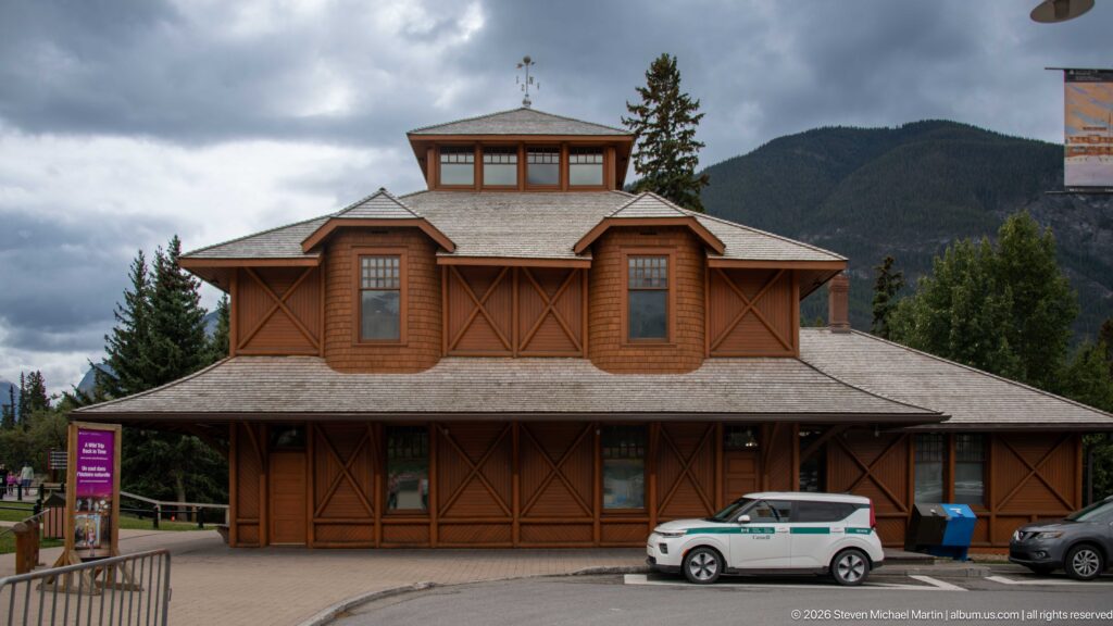 Front view of the brown timber Banff Park Museum with cross-braced walls, broad roof, clerestory, and mountains behind.