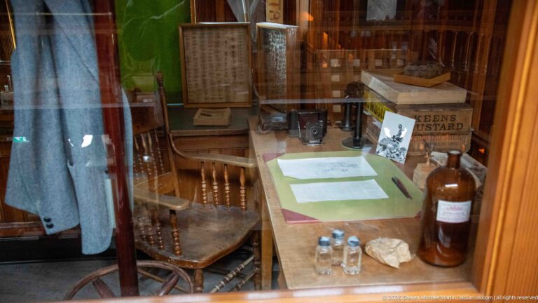 Historic curator workspace with wooden desk and chair, papers, bottles, vials, boxes, and small instruments.