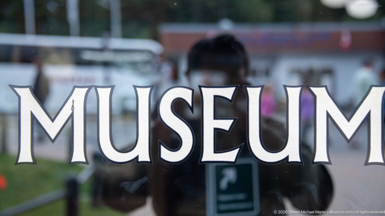 Large white MUSEUM lettering across reflective entrance glass with a person and outdoor scene visible in reflection.