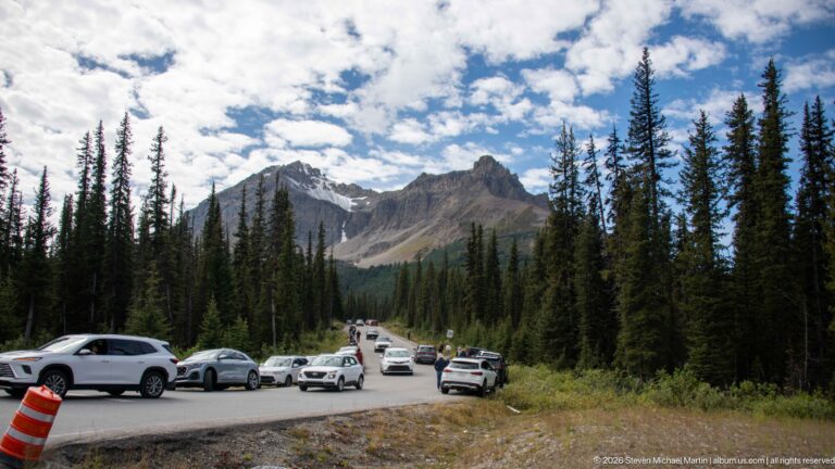 Cars parked and moving along a mountain roadside pullout, framed by tall conifers and jagged peaks under clouds.