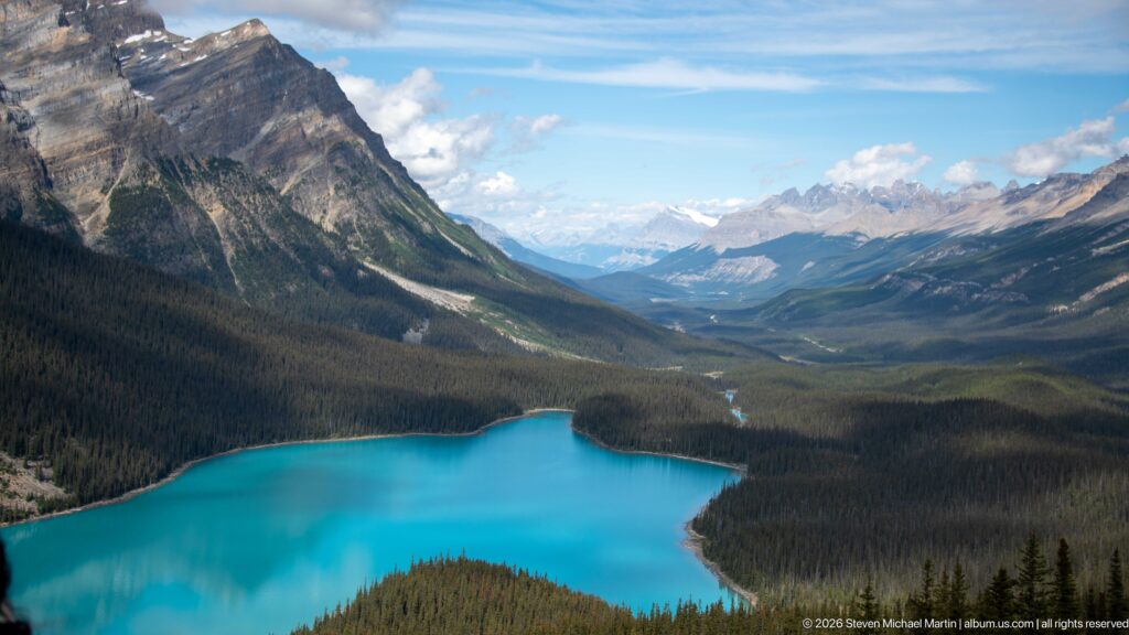 Turquoise Peyto Lake below a high viewpoint, with a forested valley and distant Canadian Rockies extending beyond the shoreline.