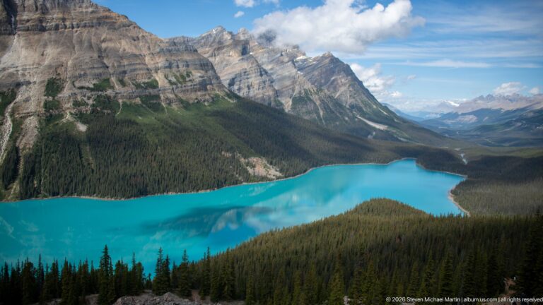 Turquoise Peyto Lake curves beneath a steep layered mountain, dark conifer forest, and distant peaks under scattered clouds.