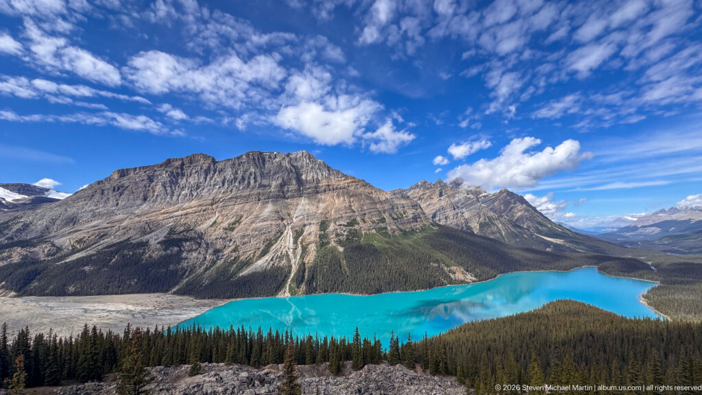 Turquoise Peyto Lake below a broad gray mountain, conifer forest, rocky foreground, and expansive cloud-streaked blue sky.