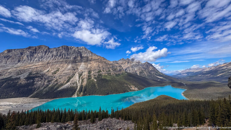 Wide view of turquoise Peyto Lake winding through forest below a massive layered mountain and distant Rocky Mountain peaks.