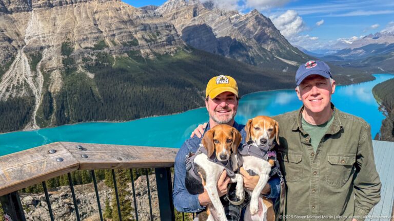 Two visitors hold beagles beside a wooden railing with turquoise Peyto Lake and mountains filling the background.