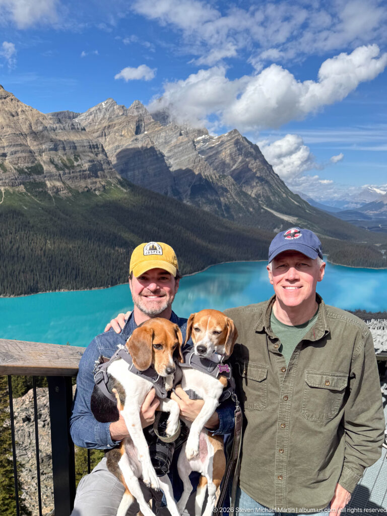 Two visitors hold beagles at a railing above bright turquoise Peyto Lake and surrounding mountain slopes.