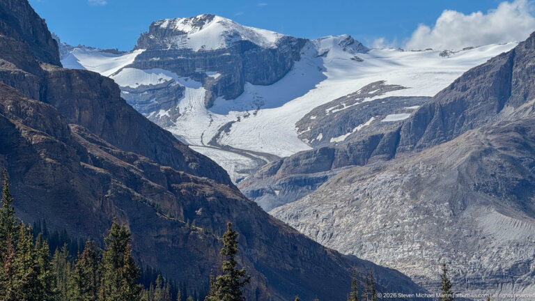 Broad white glacier descends between dark rocky walls from a high icefield above a gray debris-covered valley.