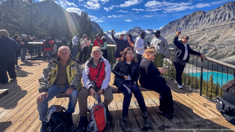 Crowded wooden overlook with seated and standing visitors above turquoise Peyto Lake and steep mountain slopes.