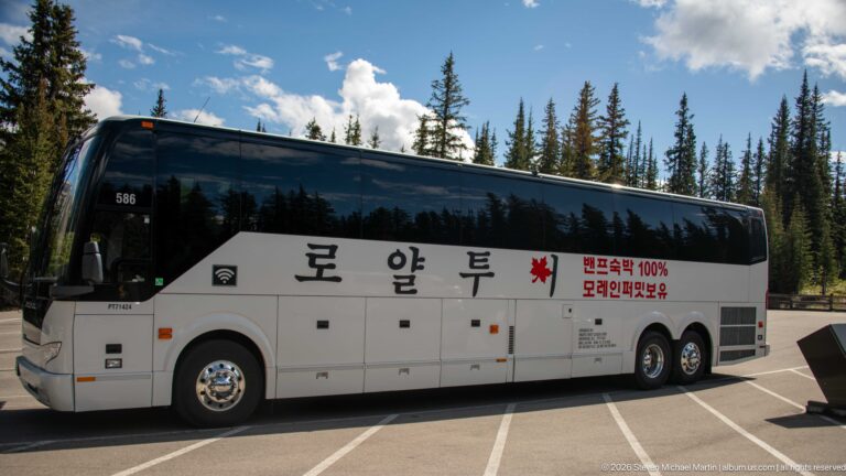 Large white tour coach with Korean lettering and a red Canadian maple leaf parked among conifers in the mountains.