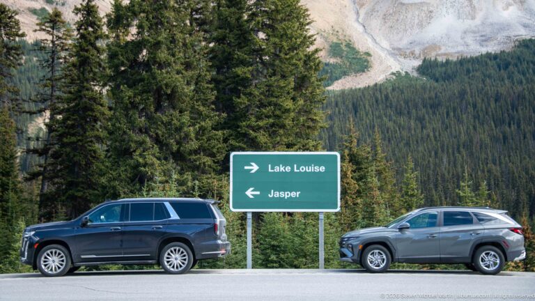 Green road sign points right to Lake Louise and left to Jasper as two SUVs pass in front of forest.