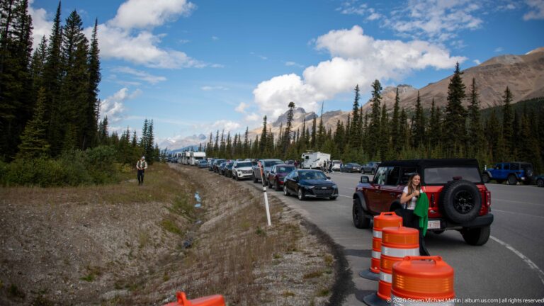 Cars parked along a mountain highway shoulder with visitors, orange traffic barrels, conifers, and rocky slopes beyond.