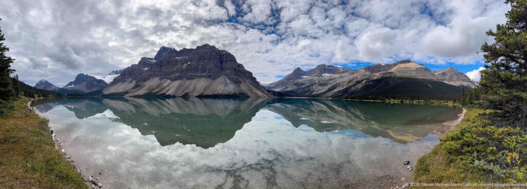 Wide view of Bow Lake reflecting several rugged mountains and clouds, framed by evergreen branches along the shore.