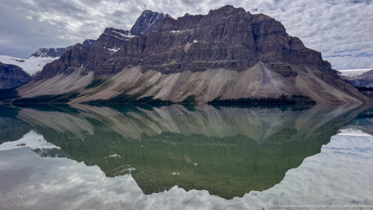 Large stratified mountain and scree slopes mirrored almost symmetrically in the green water of Bow Lake.
