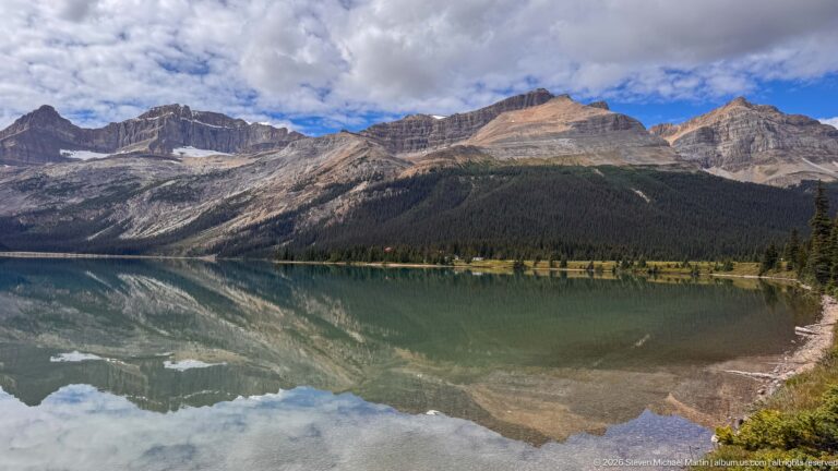 Bow Lake reflecting rugged mountains, forest, and blue sky, with a narrow vegetated shoreline across the water.