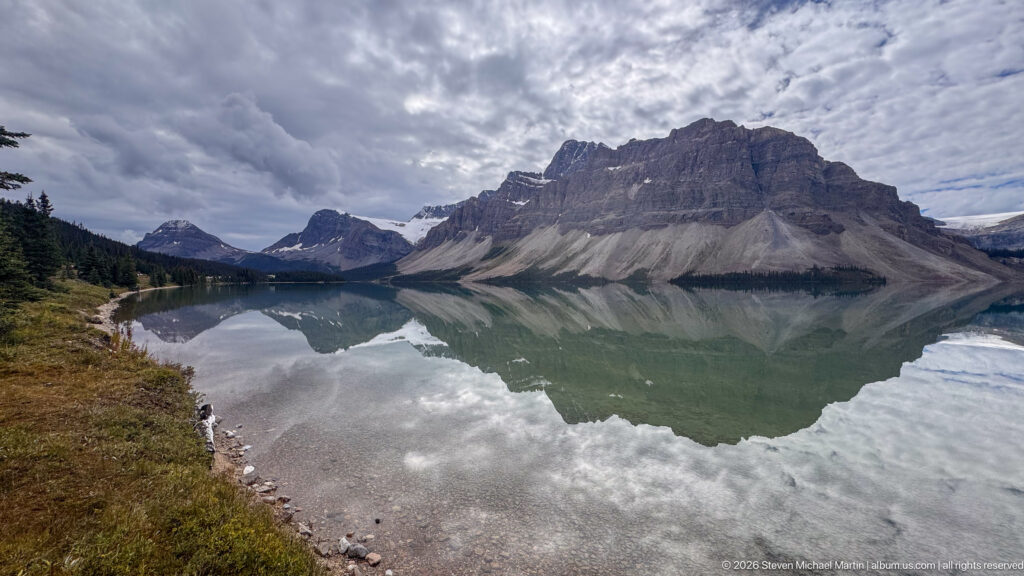 Steep layered mountain reflected in Bow Lake beside a curving gravel shore under dramatic clouds.