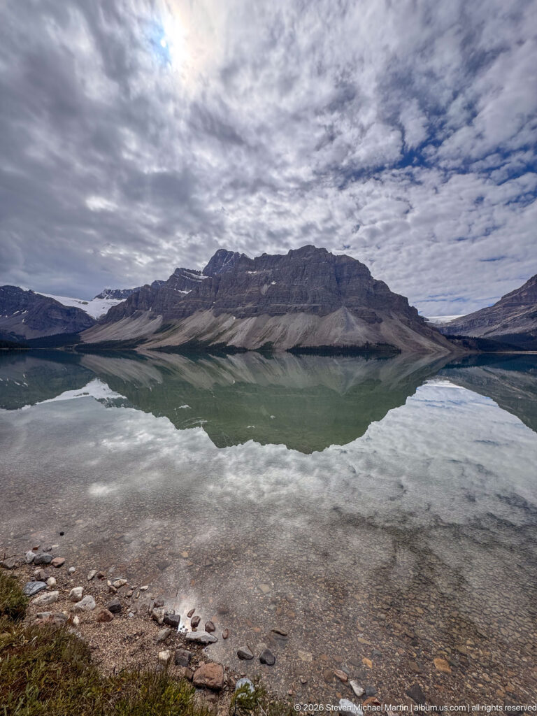 Shallow rocky shore of Bow Lake reflecting a broad mountain ridge and mottled clouds in calm water.