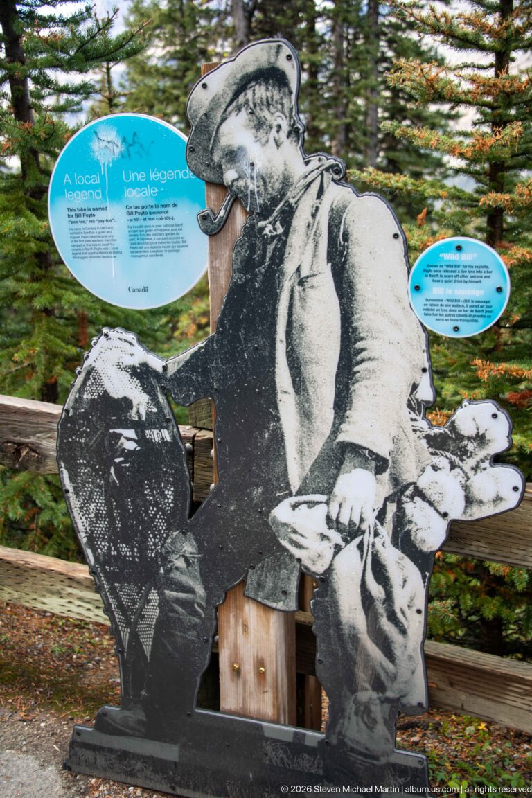 Black-and-white historic mountaineer cutout beside circular Parks Canada interpretive signs in a forested visitor area.