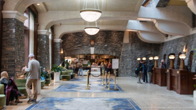 Long hotel lobby with stone walls, hanging bowl lights, reception desks, seating, guests, and a broad archway at the far end.