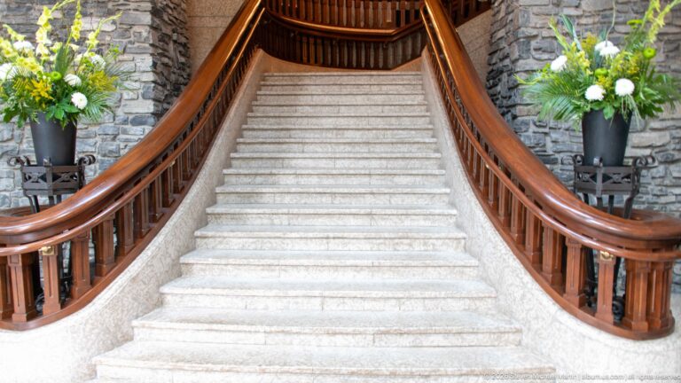 Centered view up a broad pale stone staircase with sweeping polished-wood balustrades, dark stone walls, and floral arrangements on both sides.