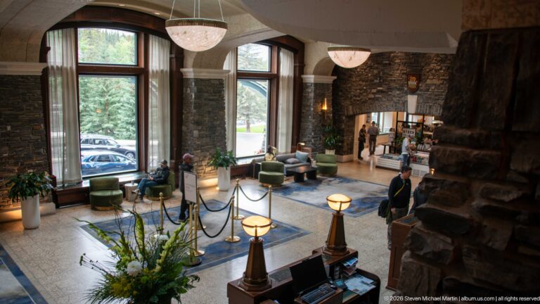 Elevated lobby view with tall windows, stone walls, green chairs, blue rugs, hanging lights, floral arrangement, and guests.
