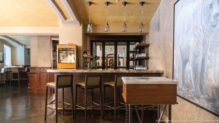 Modern hotel bar with dark counter, high stools, wood cabinetry, beverage equipment, pendant lights, and a large pale abstract artwork.