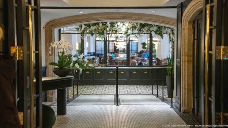 Arched interior doorway with glass doors, patterned floor, orchid arrangement, and a bright dining room with greenery and guests beyond.