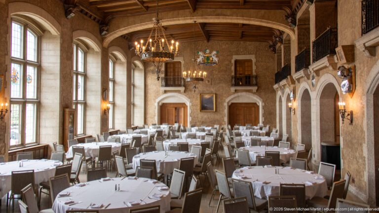 Long formal dining hall with tall windows, round tables, chandeliers, timber ceiling, arched wall bays, and an upper gallery.