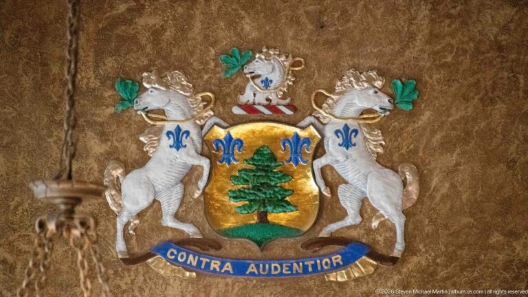 Decorative coat of arms with two white animal supporters, gold shield, green tree, blue fleur-de-lis, and Contra Audentior motto.