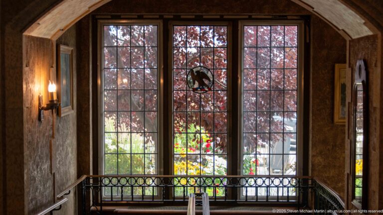 Tall multi-pane windows in an arched opening overlook red foliage and bright garden flowers behind a dark interior railing.