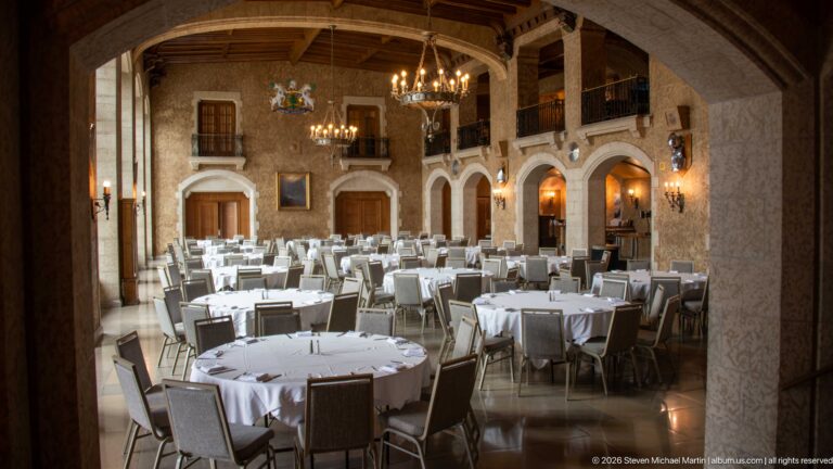 Large formal dining hall with white-clothed round tables, timber ceiling, chandeliers, arches, balconies, and heraldic wall decoration.