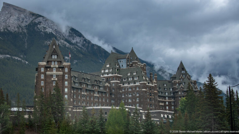 Banff Springs Hotel towers above conifers with steep roofs and dark stone walls, backed by cloud-wrapped Rocky Mountain slopes.