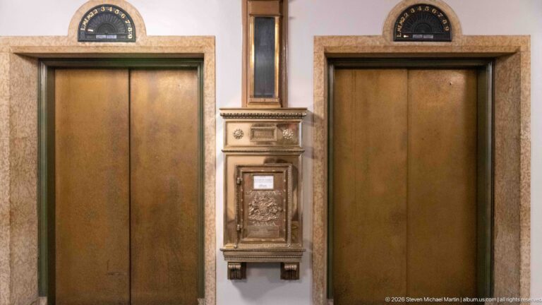 Two brass-toned elevator doors flank an ornate wall-mounted call station, with an arched floor indicator above the right door.