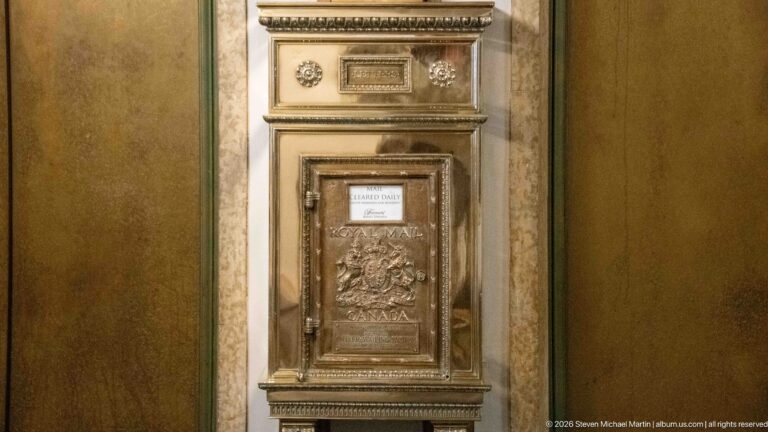 Tall ornate metal letter box with molded decoration and relief panel mounted against an interior wall.