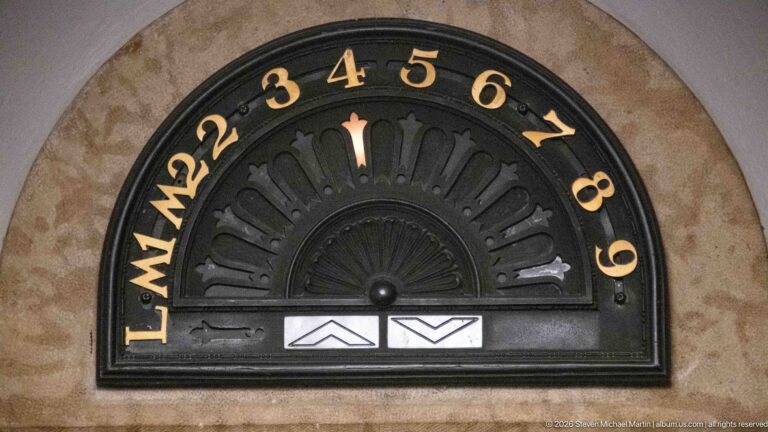 Semicircular black metal elevator floor indicator with gold numbers, central pointer, lettered positions, and up-and-down arrows.