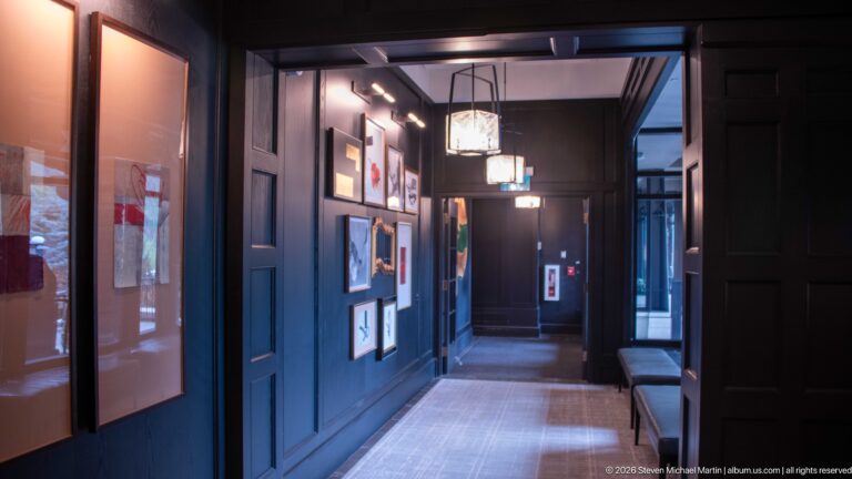 Long dark-paneled hotel corridor with framed artwork, overhead lanterns, a bench, and a pale runner leading toward a doorway.