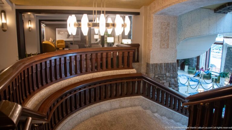 Curving staircase with dark wooden balustrade and pale stone steps beside an upper landing lit by clustered pendant lights.