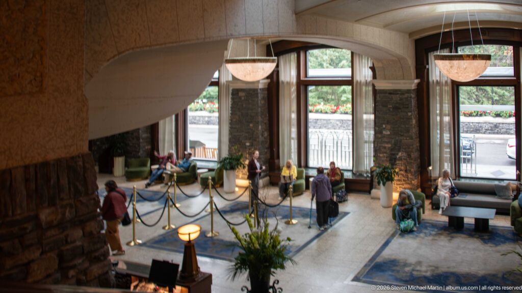 Elevated view into a hotel lobby with tall windows, stone piers, blue carpet, hanging lights, seating, and several guests.