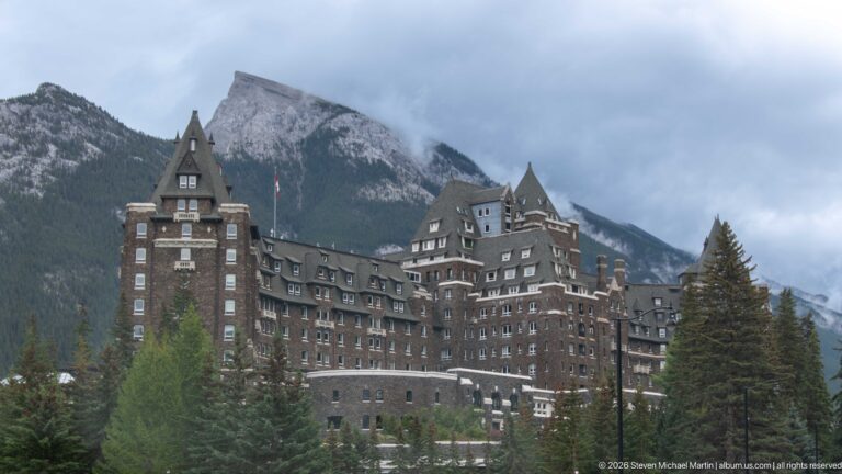 Dark stone towers and dormered roofs of Banff Springs Hotel rise above evergreens beneath a misty mountain slope.