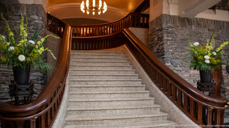 Broad pale stone staircase with curved polished-wood railings, stone walls, floral arrangements, and warm ceiling lights.