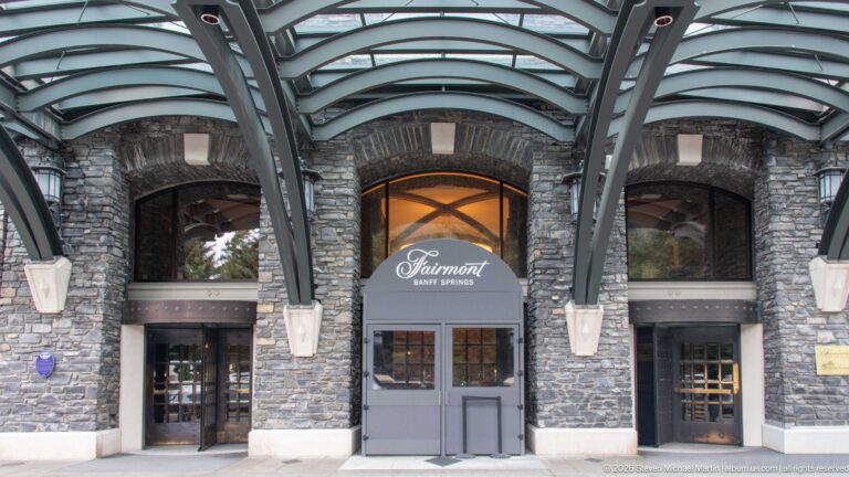 Three arched stone entrance bays beneath a curved glass canopy, with Fairmont Banff Springs lettering above the central doors.