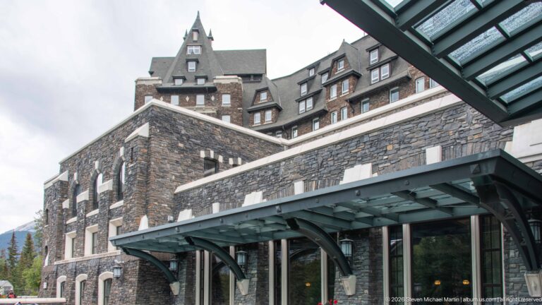 Stone hotel façade with arched openings, a glass-and-metal entrance canopy, dormers, and steep gables rising above.