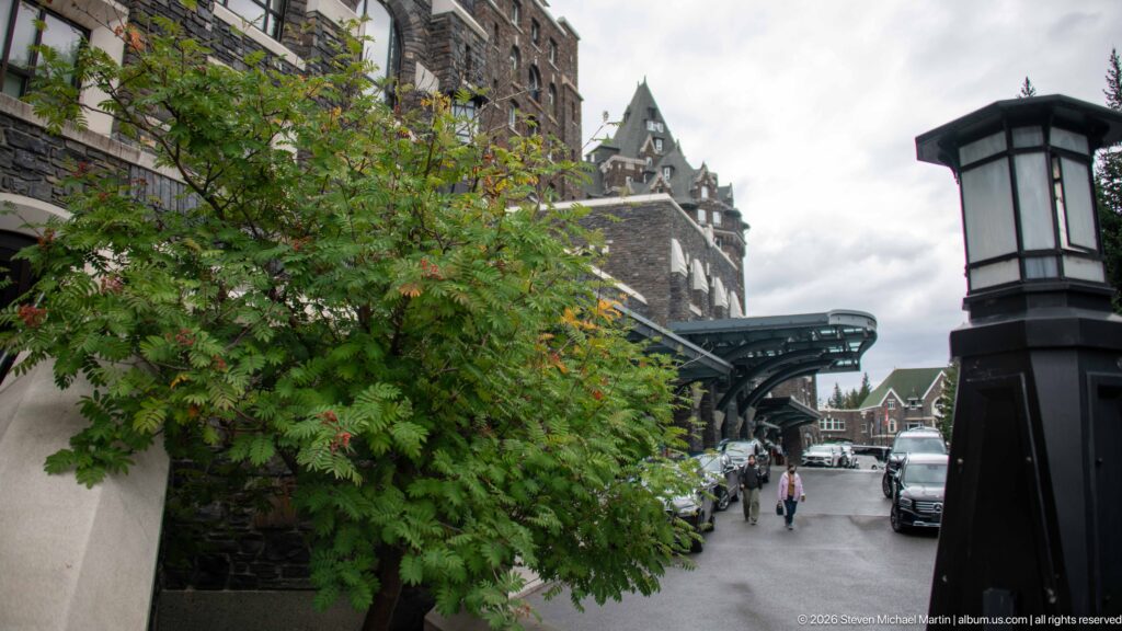 Tree-lined hotel approach with vehicles, pedestrians, a tall lantern, and the Banff Springs Hotel rising beyond a covered entrance.
