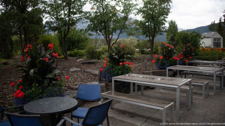 Metal tables, benches, and chairs on gravel among tall red flowers and trees, with mountains and a small building beyond.