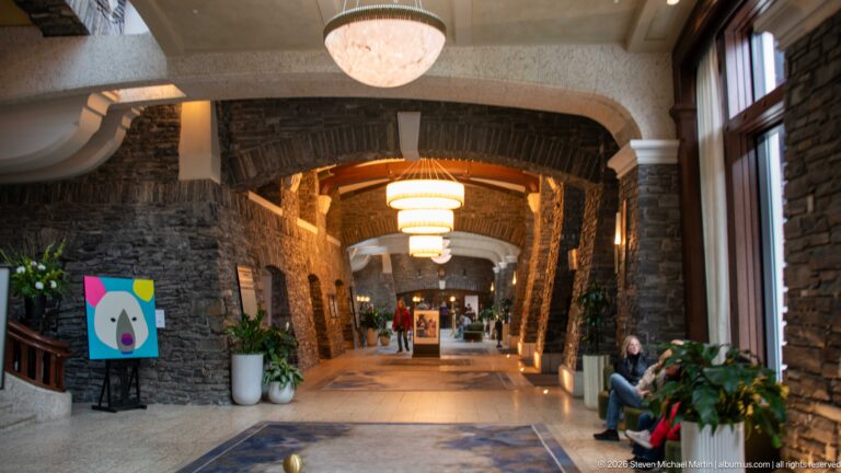 Long Banff Springs Hotel corridor with dark stone arches, pale vaulted ceiling, chandeliers, large windows, artwork, and plants.