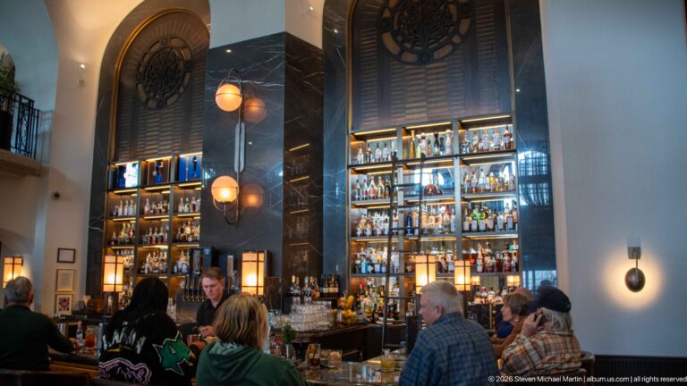 Busy Rundle Bar with seated guests below tall illuminated bottle shelves, dark wall panels, and glowing pendant lights.