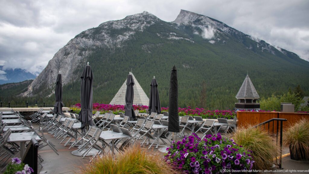 Rows of metal terrace chairs and closed umbrellas with purple flowers, set against a steep forested Rocky Mountain peak.