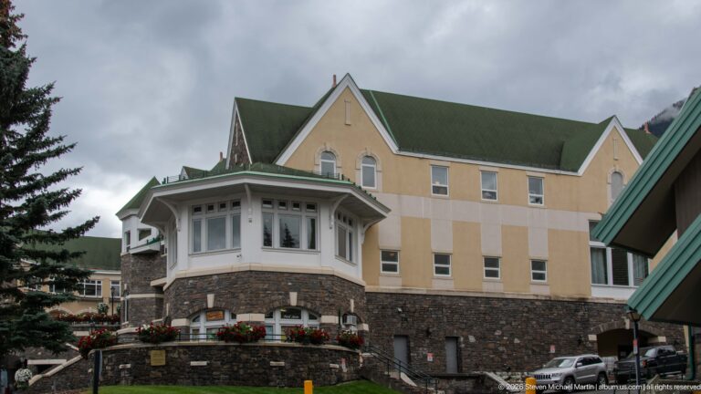 Pale stucco hotel wing on a dark stone base with green gabled roof, regular windows, and a projecting enclosed bay.